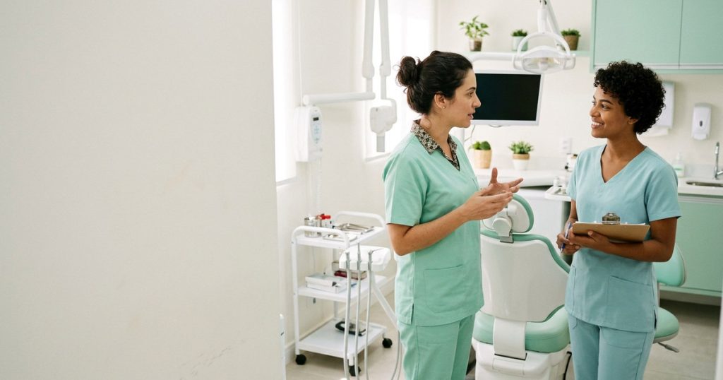 Dentista e auxiliar conversam ao lado da cadeira em um consultório odontológico